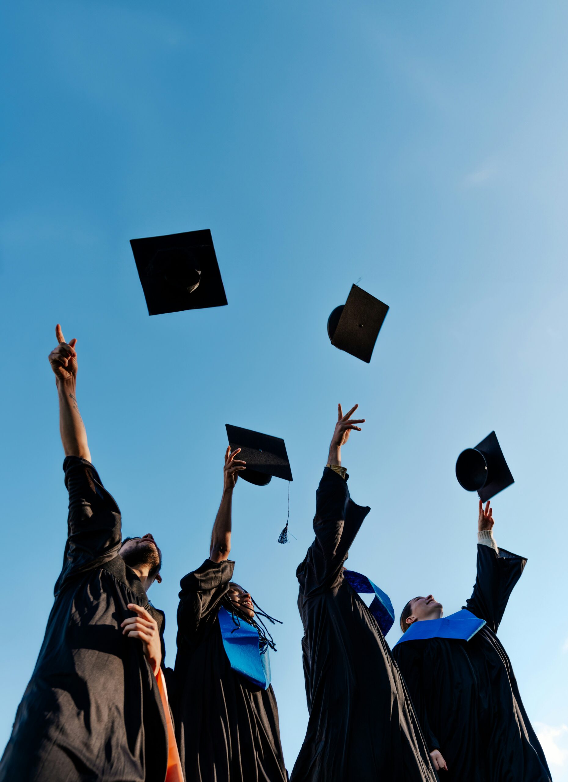 Low-angle view of four diverse college graduates in black gowns throwing their mortarboard caps into a clear blue sky, celebrating their graduation.