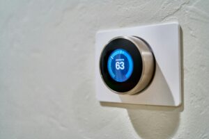 A close-up, angled shot of a modern, circular smart thermostat mounted on a textured white wall. The sleek device features a metallic outer ring and a glowing blue digital display that reads "HEAT SET TO 63".
